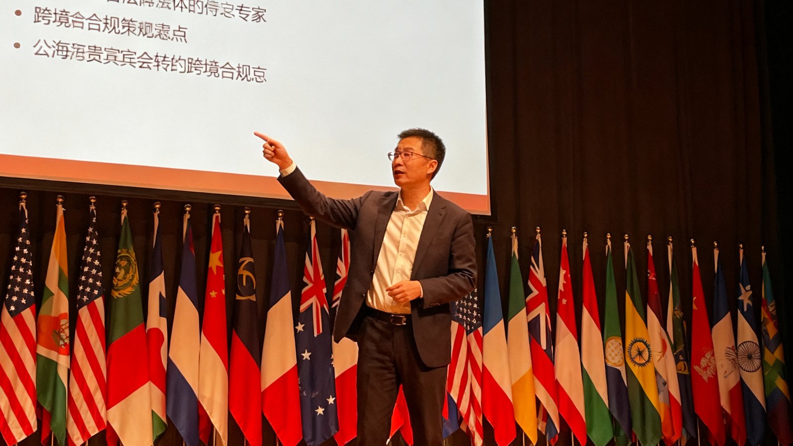 公海贵宾会特约跨境合规专家何志远在国际论坛上解读政策，背景为多国国旗排列的舞台，他正指向屏幕上的法规要点